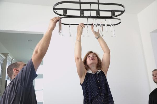 A home stager hangs a small, black chandelier in the middle of a white room with help from another assistant.