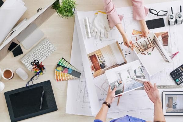 From directly above, we see two interior designers at a desk across blueprints, color samples, a Mac desktop computer, a tablet, a calculator, a small green plant, and pictures of the interior of a house they're currently working on.