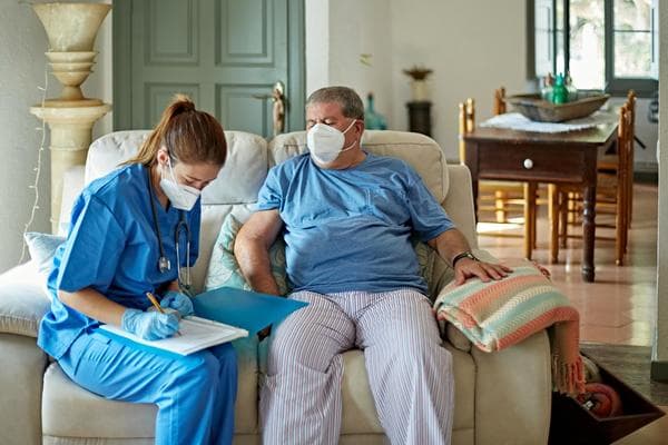 A home healthcare provider sits next to her male patient on a couch in the patient's home and takes down notes.