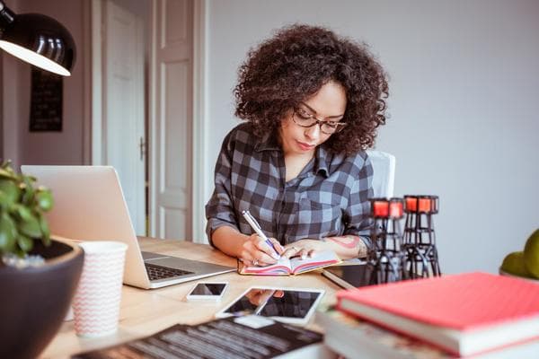 A home organizer sits at her desk in front of her laptop as she writes notes down in a little notebook. On the desk, we see a pink book, a tablet, a smartphone, a green plant, and a black lamp.