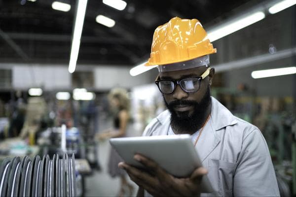 An engineer wearing a yellow hard hat and holding a digital tablet stands in a warehouse, examining the contents of the tablet.